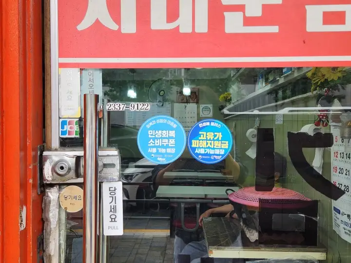 매장 출입문에 지난해 '민생회복소비쿠폰'과 올해 '고유가 피해지원금' 사용 가능 스티커가 함께 붙어 있다. 정부 소비지원 정책이 지난해부터 이어지고 있음을 보여준다.